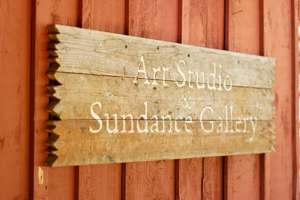 A weathered wooden sign with white text reading "Art Studio & Sundance Gallery" mounted on a red wooden wall.