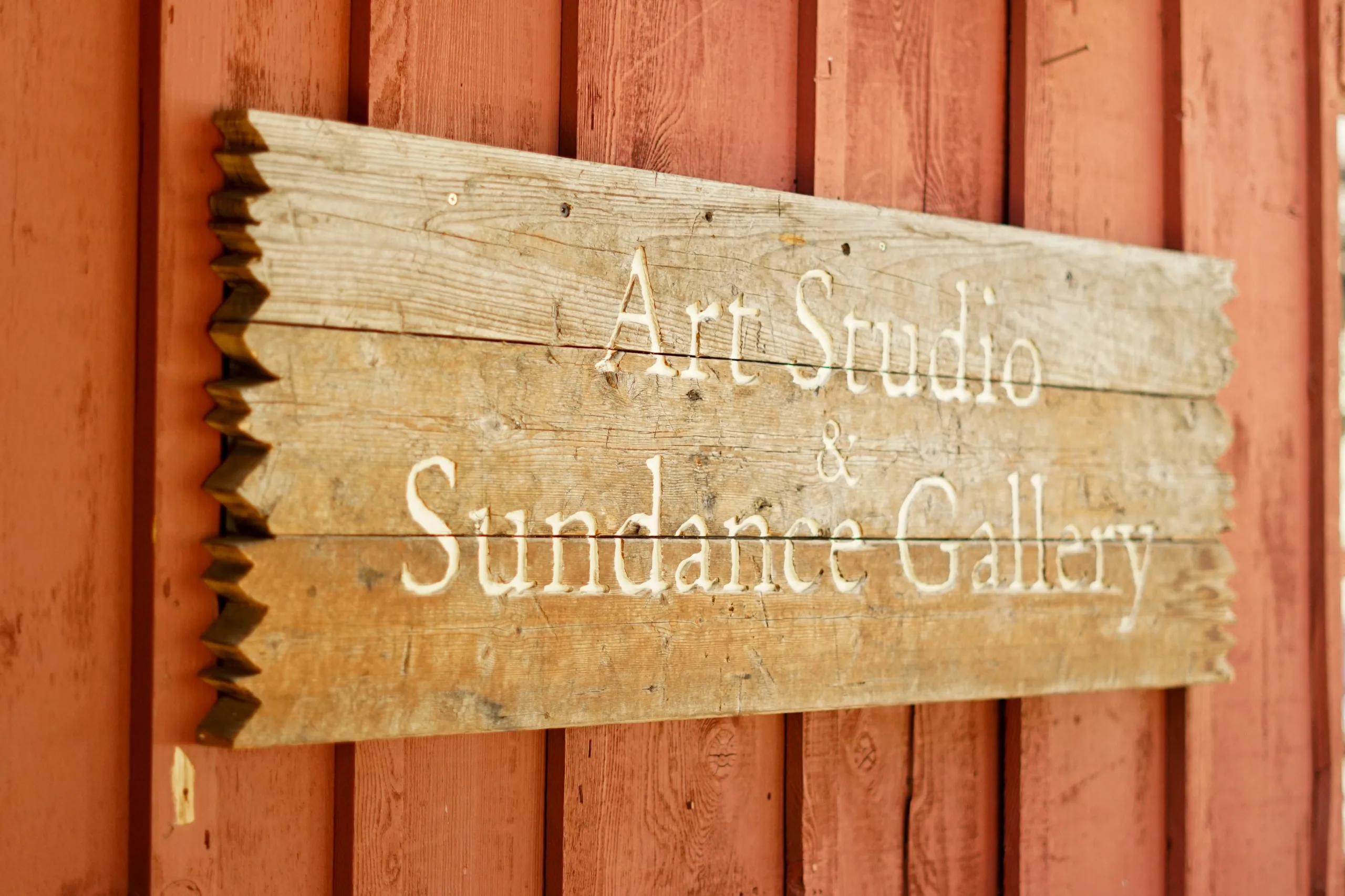 A weathered wooden sign with white text reading "Art Studio & Sundance Gallery" mounted on a red wooden wall.