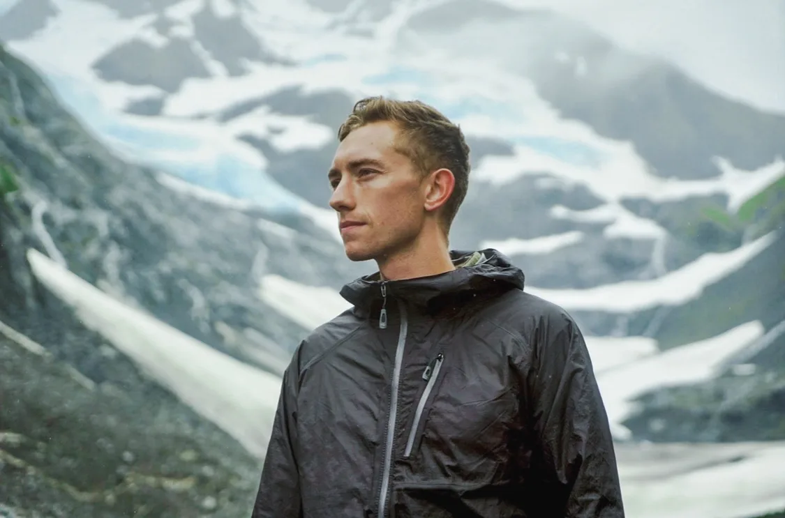 A person wearing a black rain jacket stands outdoors in front of a rocky, snow-covered mountain landscape, looking to the side with a calm expression.
