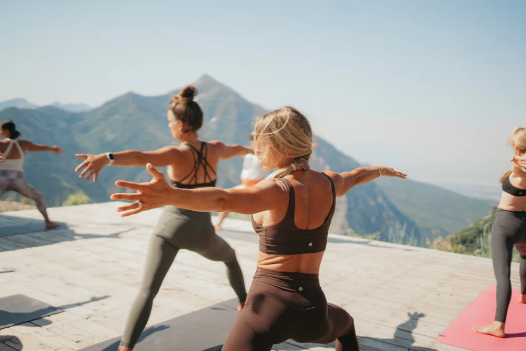 People practicing yoga on a wooden platform with a scenic mountain view. They are doing poses with arms extended, wearing sportswear. The sky is clear and the atmosphere is serene.