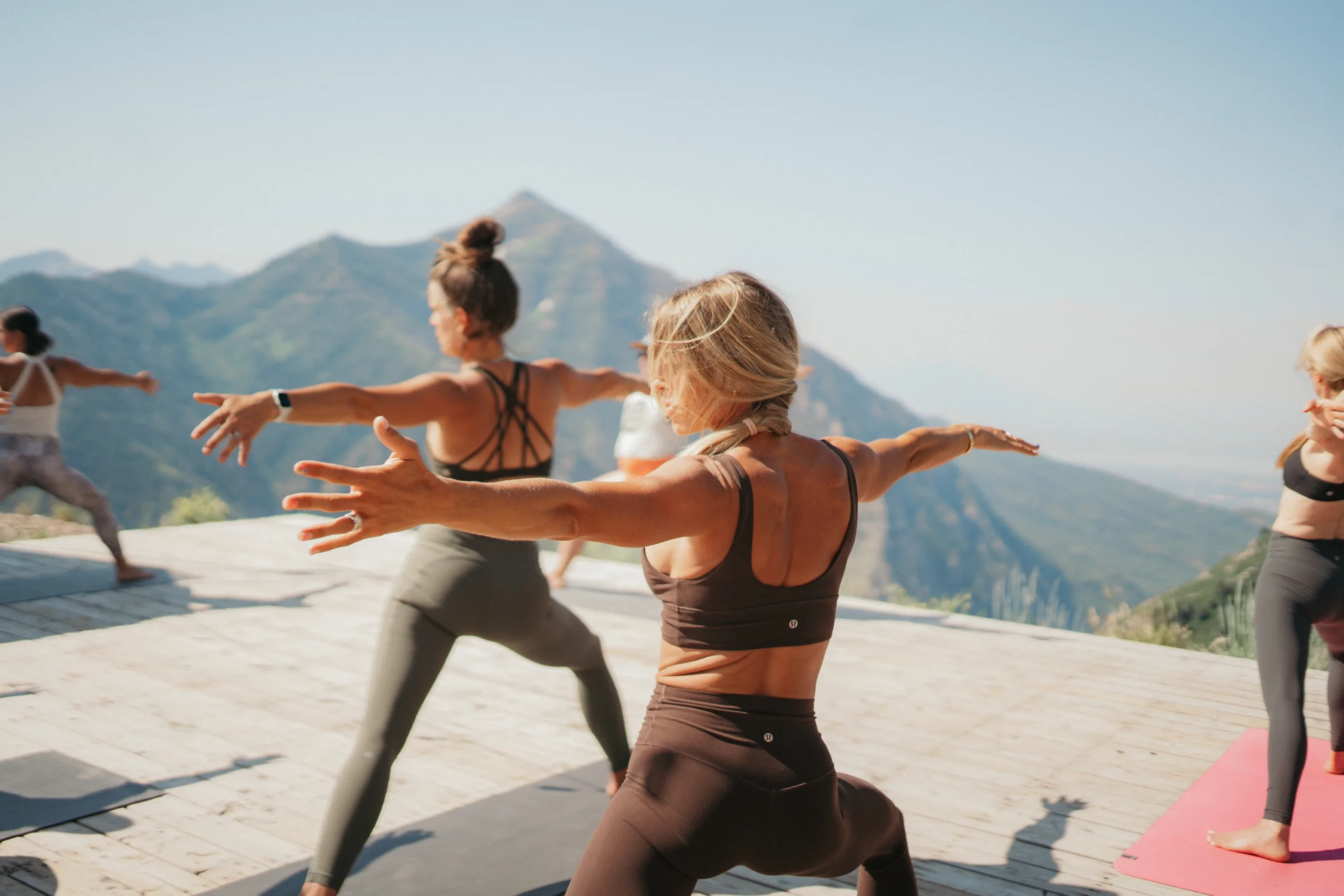 People practicing yoga on a wooden platform with a scenic mountain view. They are doing poses with arms extended, wearing sportswear. The sky is clear and the atmosphere is serene.