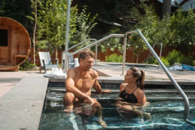 A man and woman in swimwear relax and talk together in an outdoor hot tub surrounded by trees and patio furniture on a sunny day.