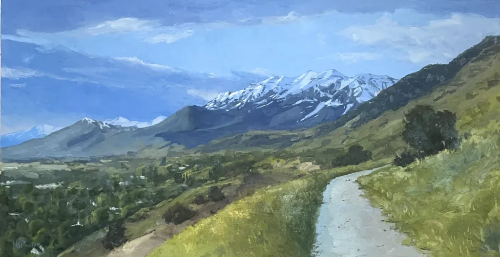 A winding path leads through grassy hills toward distant snow-capped mountains under a blue sky with scattered clouds. Trees and greenery fill the landscape.