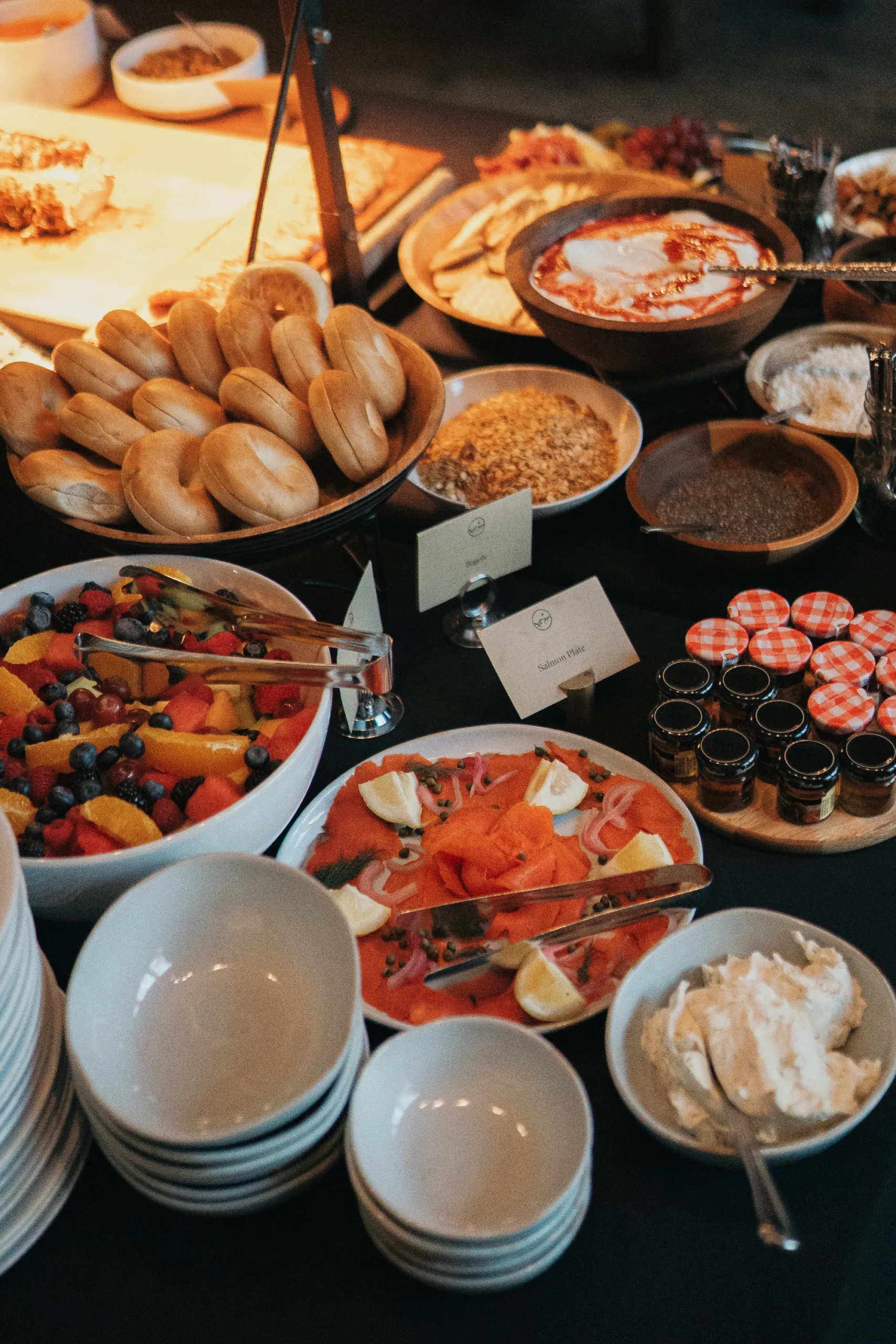 A breakfast buffet featuring bagels, smoked salmon with lemons, bowls of mixed fruit, small jars of preserves, and bowls of granola and yogurt. Plates and serving utensils are arranged around the spread.