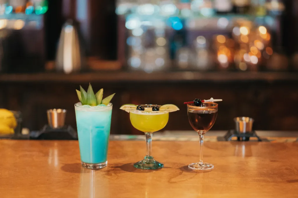 Three cocktails are lined up on a bar counter. The first is a blue drink in a highball glass garnished with pineapple leaves. The second is a green drink in a coupe glass with limes and berries. The third is a dark drink in a martini glass with a cherry.