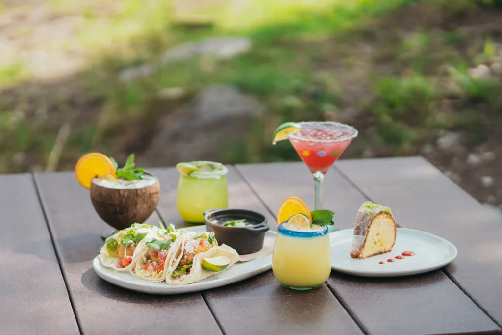 A wooden table outside with various foods and drinks. There are three tacos on a plate with lime wedges, a slice of cake, two cocktails, a margarita in a glass with a lime wedge, and a coconut drink garnished with orange and mint.