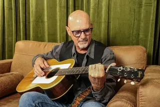 A bald man with glasses sits on a brown couch, playing an acoustic guitar. He wears a dark shirt, jeans, and a black vest, with a green curtain in the background.