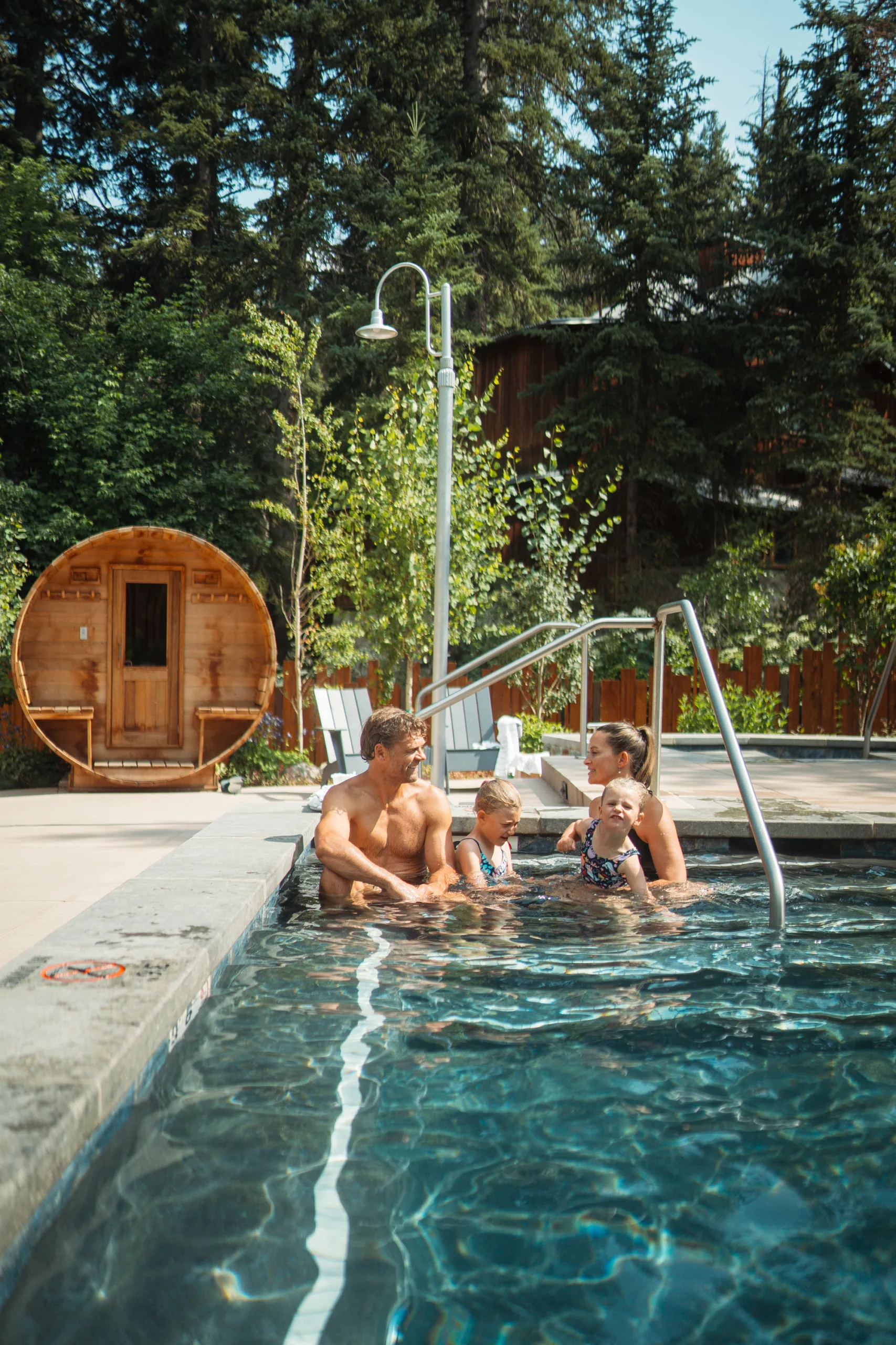 A family of four, two adults and two children, enjoy time together in an outdoor pool surrounded by trees. A wooden sauna sits in the background on a sunny day.