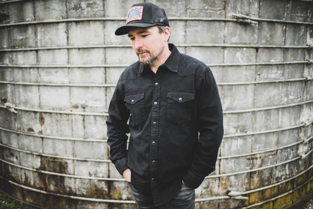 A man wearing a black button-up shirt and a baseball cap stands with his hands in his pockets in front of a large, weathered metal tank. He looks down, appearing thoughtful or contemplative.