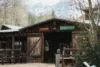 A rustic wooden general store with open doors and benches outside, surrounded by trees, with snow-capped mountains visible in the background.