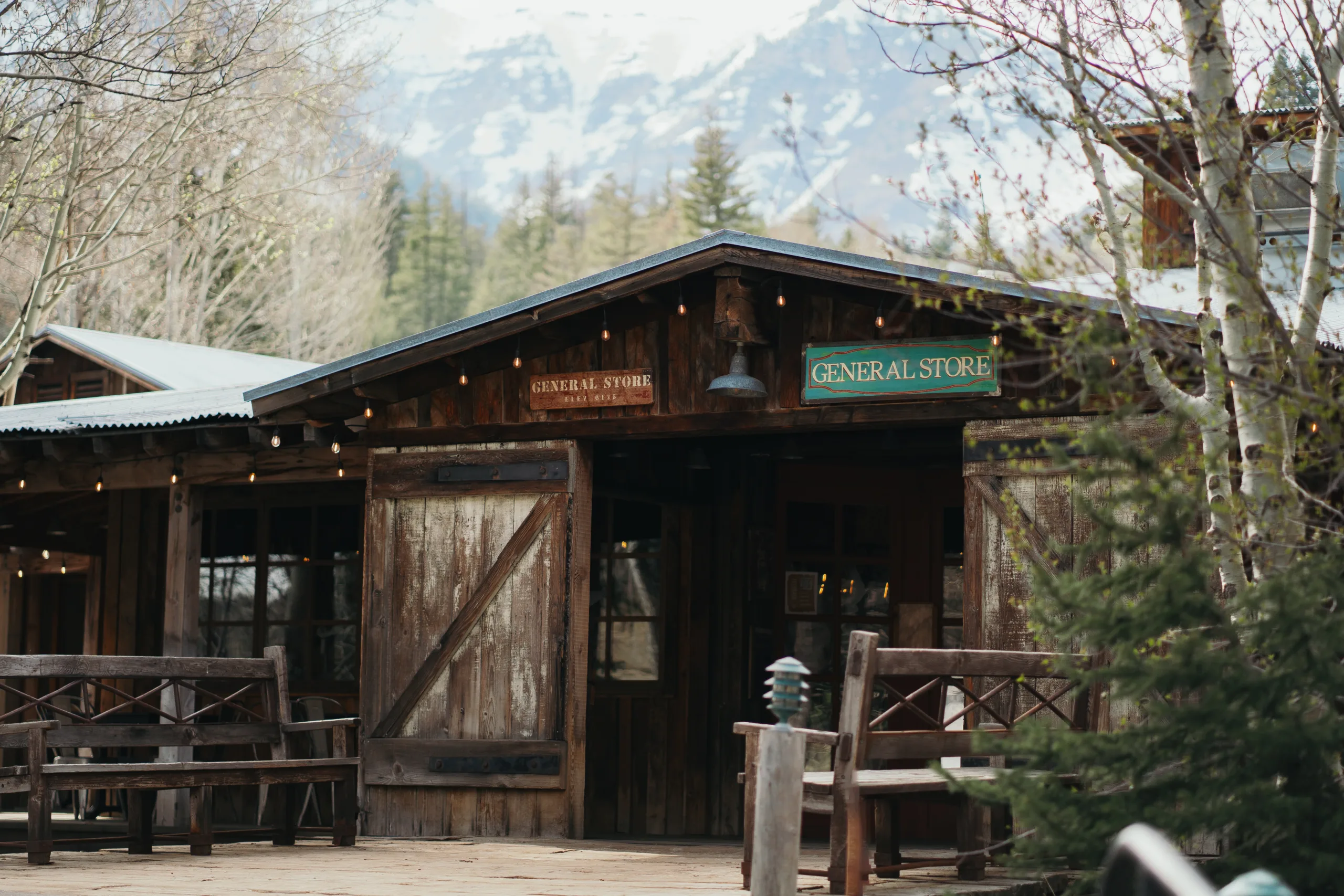 A rustic wooden general store with open doors and benches outside, surrounded by trees, with snow-capped mountains visible in the background.