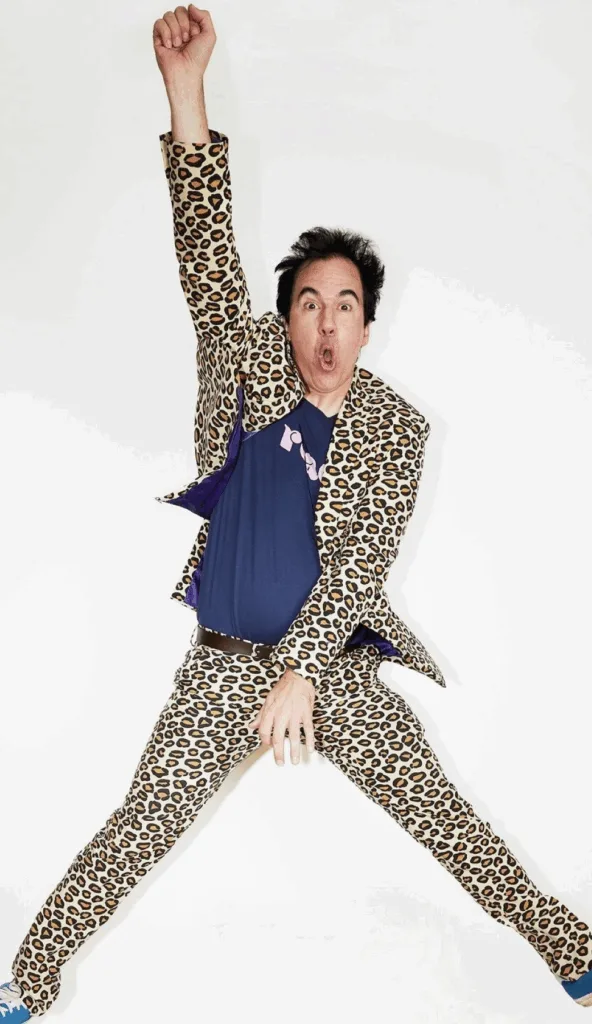 A man wearing a leopard-print suit and dark t-shirt jumps energetically against a white background, raising one arm above his head and spreading his legs wide.