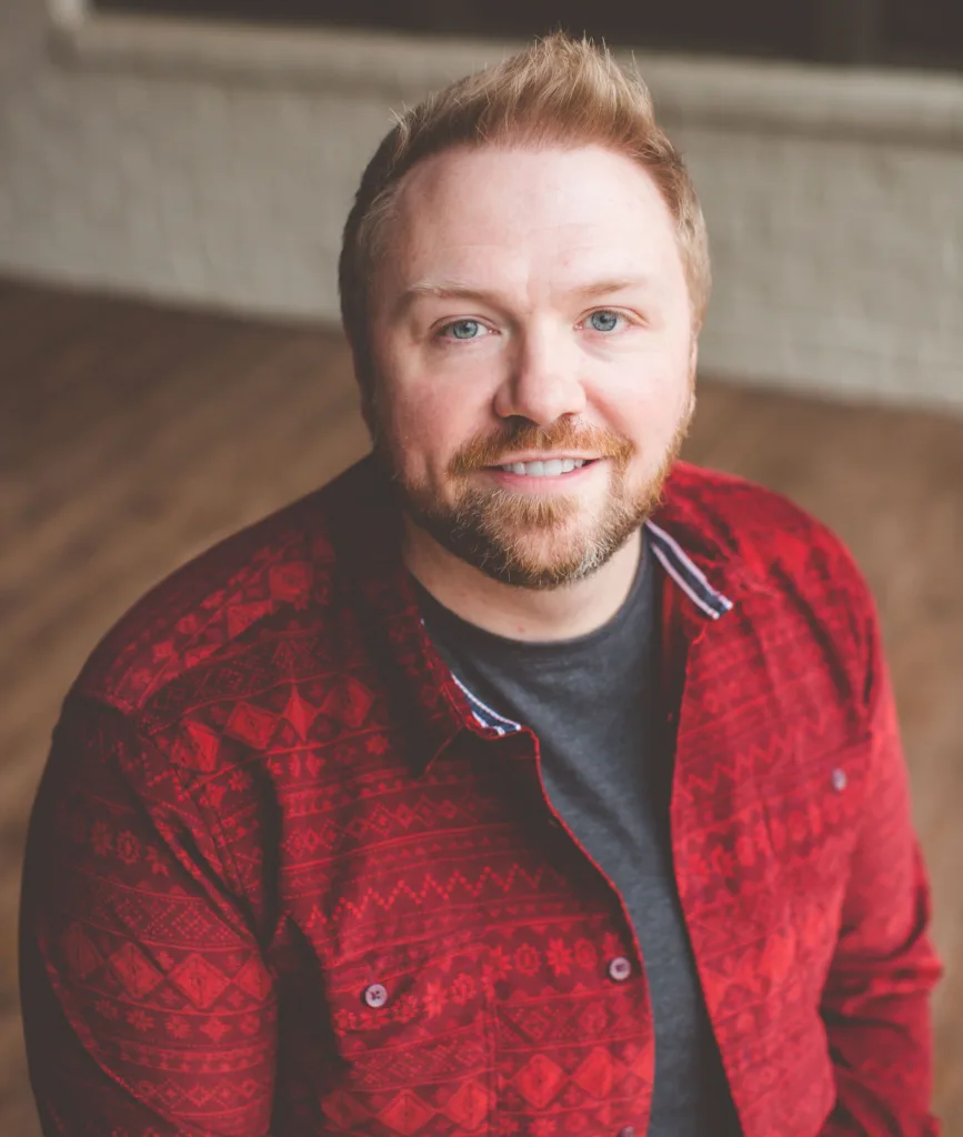 A smiling man with light hair and a short beard, wearing a red patterned button-up shirt over a dark t-shirt, sits indoors on a wooden floor with a blurred background.