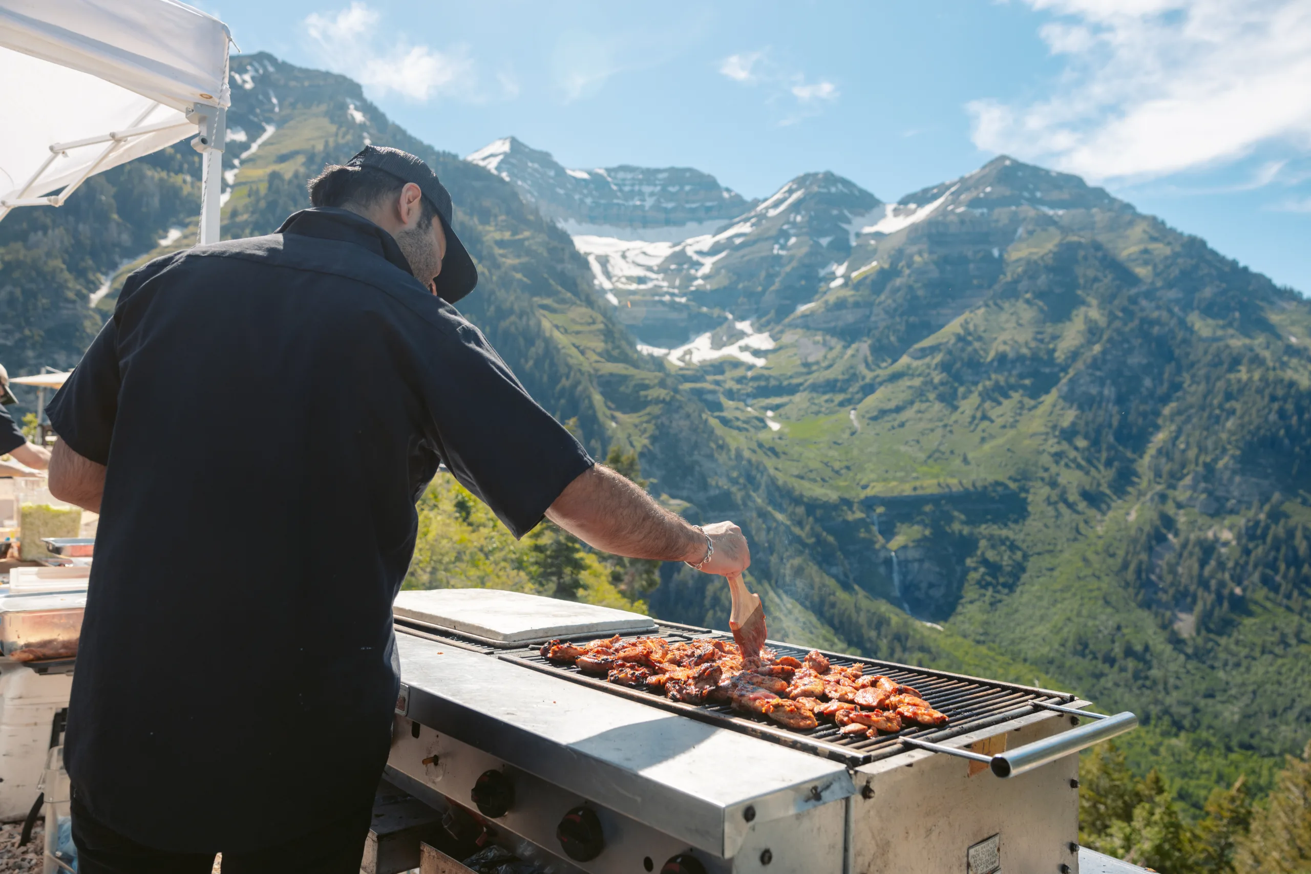A person grills meat on a barbecue outdoors with scenic, green mountains and patches of snow in the background under a clear, sunny sky.