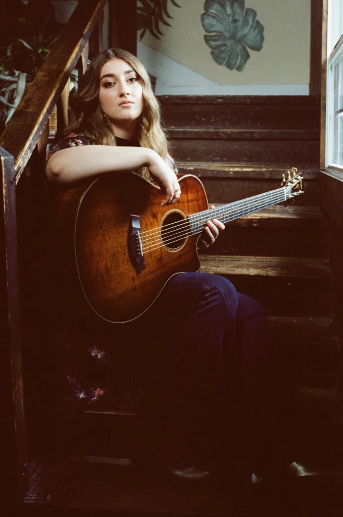 A young woman with long hair sits on wooden stairs, holding an acoustic guitar. She gazes calmly at the camera. Green plants and natural light from a window create a cozy, artistic atmosphere.