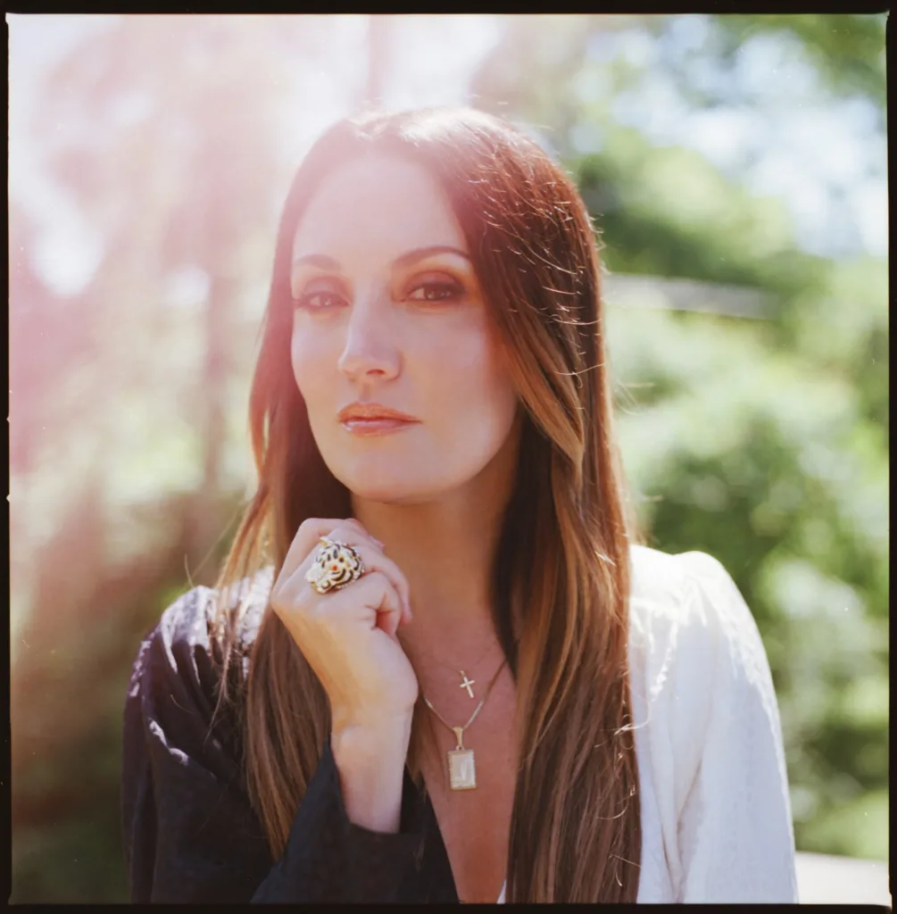 A woman with long brown hair stands outdoors, wearing a dark top and white jacket. She gazes forward, holding her hand to her chin, and wears gold jewelry. Sunlight and greenery are visible in the blurred background.