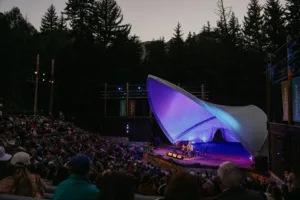 An outdoor amphitheater with a large crowd watching musicians perform on a lit stage under a white, shell-shaped canopy at dusk, surrounded by tall pine trees.