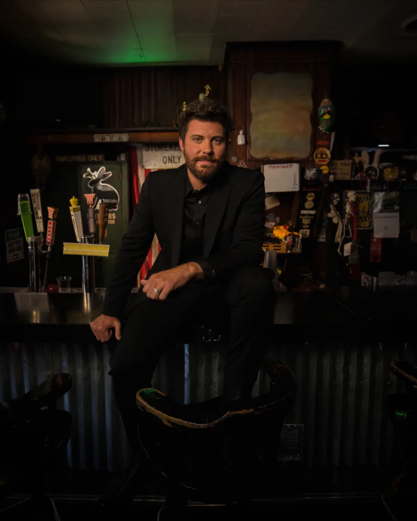 A man with a beard, dressed in a black suit, sits on the counter of a dimly lit bar, surrounded by colorful beer tap handles and eclectic bar decor.