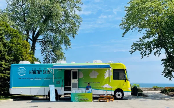 A colorful mobile clinic with "Destination Healthy Skin" written on the side is parked outdoors near trees and water. A person sits at a table with informational materials set up in front of the vehicle.