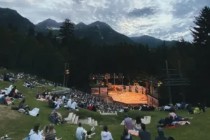 A Summer Theatre performance unfolds at dusk in a mountain setting, with audience members seated on a grassy hillside, surrounded by trees and distant peaks beneath a cloudy sky.