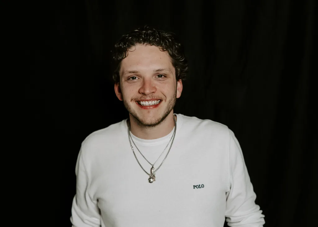 A young man with curly hair smiles at the camera, wearing a white long-sleeve shirt with "POLO" on it and two silver necklaces. The background is black.