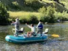 Three people wearing hats and life jackets are fishing from an inflatable blue raft on a calm river, surrounded by greenery and sunlight.